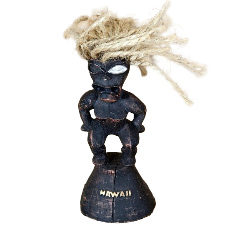 A small carved black Hawaiian-style figure with a natural fiber hairpiece stands on a round base engraved with the word “HAWAII,” 