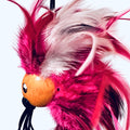 A close-up of a colorful bird-shaped toy with bright pink and white feathers, a round orange beak, and black hanging strings against a plain light background.