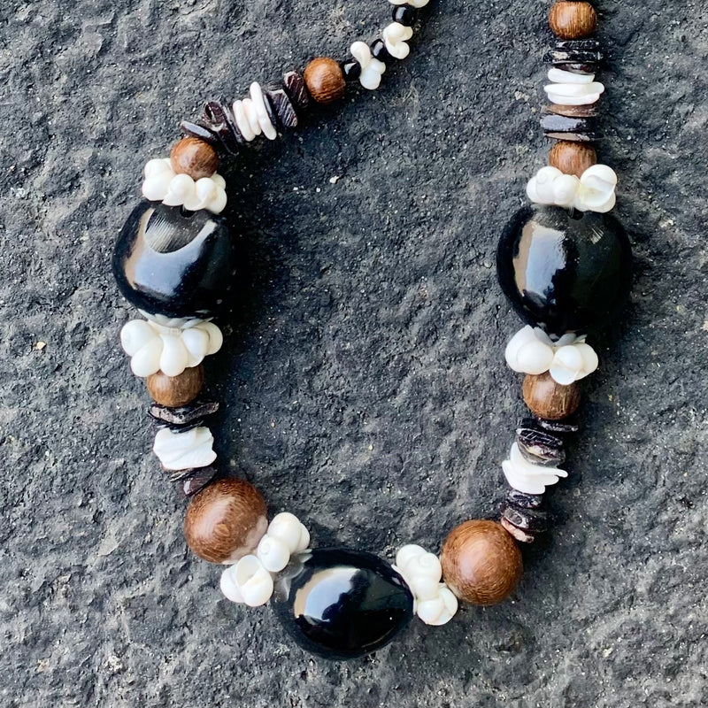 Handmade Hawaiian-style lei necklace with black kukui nuts, brown wood beads, and white mongo shell accents on a textured stone background
