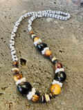 Handmade Hawaiian-style lei necklace with black kukui nuts, light brown wood beads, and white mongo shell accents on a textured stone background