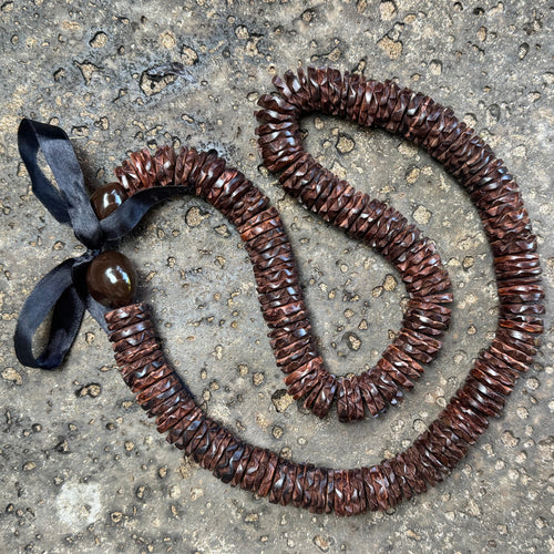 A brown ccocnut wood necklace made of many small overlapping pieces, with a black ribbon tied at one end and a round brown kukui nut, aid on a rough gray concrete surface.