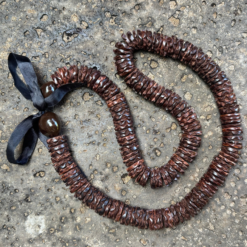 A brown ccocnut wood necklace made of many small overlapping pieces, with a black ribbon tied at one end and a round brown kukui nut, aid on a rough gray concrete surface.