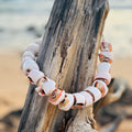 Necklace with shell beads hanging on a piece of driftwood by a beach.

