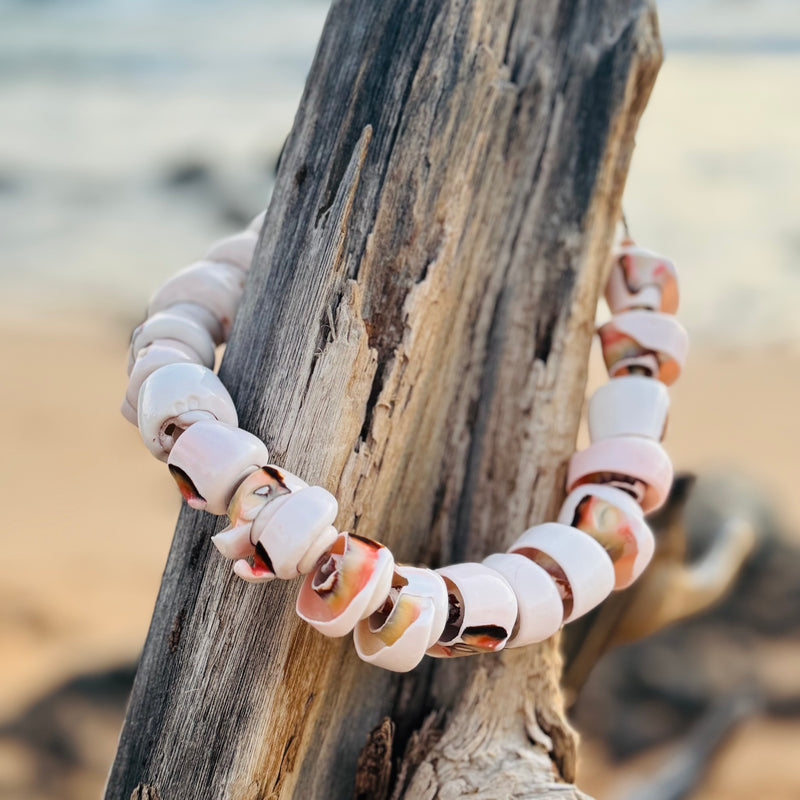 Necklace with shell beads hanging on a piece of driftwood by a beach.

