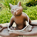 Bronze statue of a cat in a meditative pose with a blurred green garden background wearing a puka shell necklace