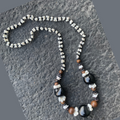 Handmade Hawaiian-style lei necklace with black kukui nuts, brown wood beads, and white mongo shell accents on a textured stone background