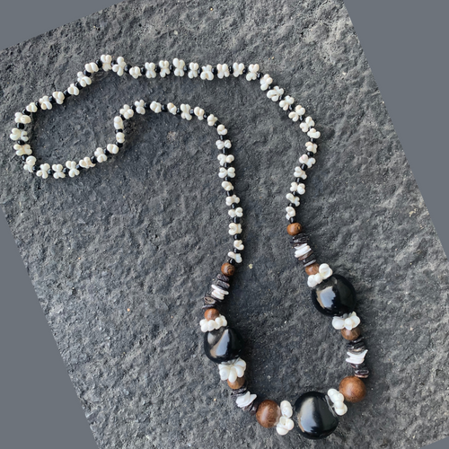 Handmade Hawaiian-style lei necklace with black kukui nuts, brown wood beads, and white mongo shell accents on a textured stone background